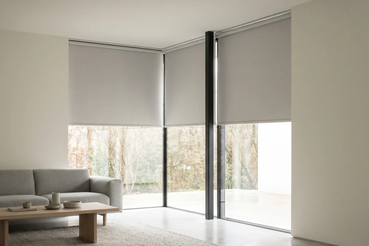 Roller blinds lowered to the same height across a glass corner window, meeting at the slim corner post