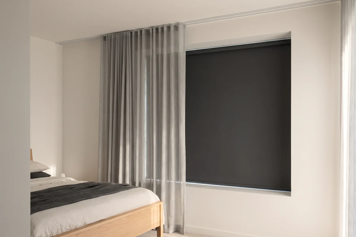 A bedroom window with a blockout blind behind a floor-length sheer curtain