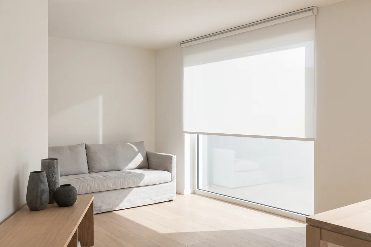A white roller blind half lowered over a wide living room window, a linen sofa beside it