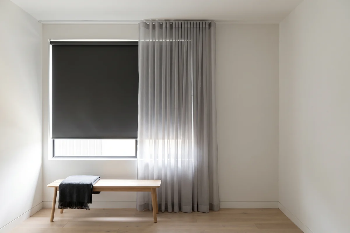 A blockout roller blind lowered behind a sheer curtain hanging from runners on a ceiling track