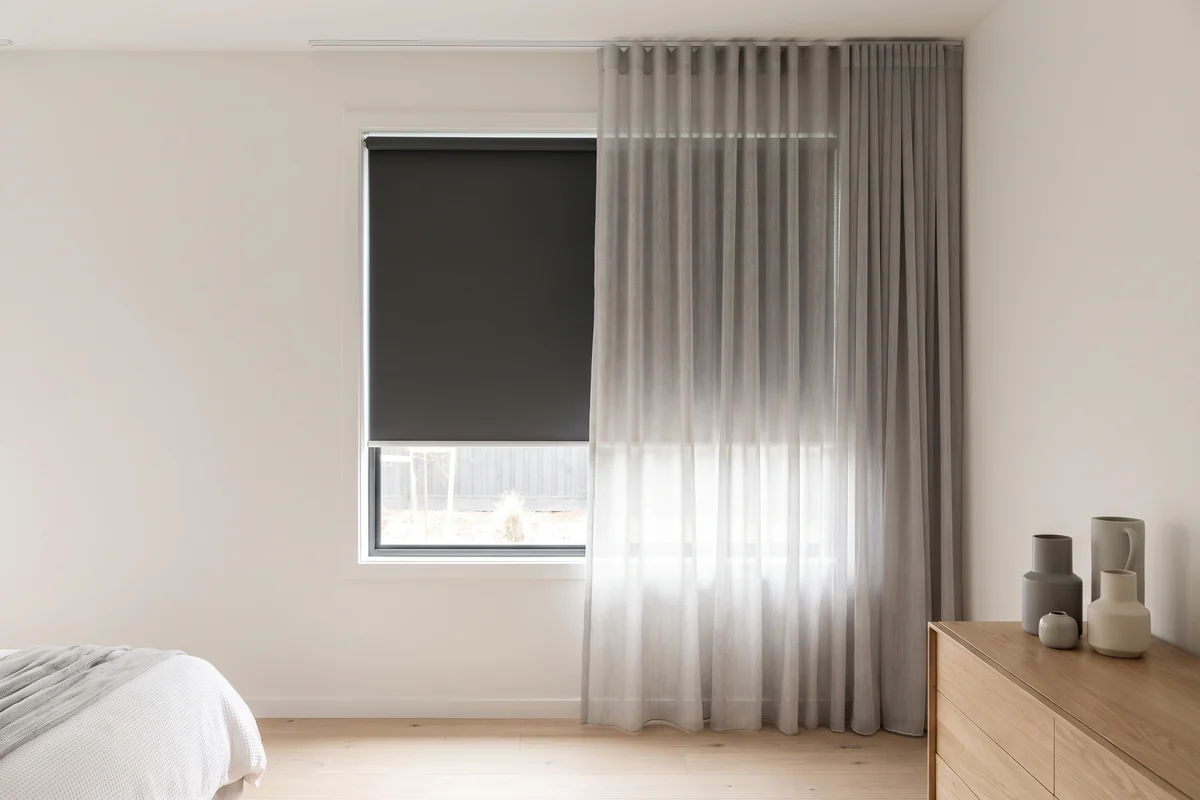 A bedroom window with a blockout blind and a sheer curtain in front of it