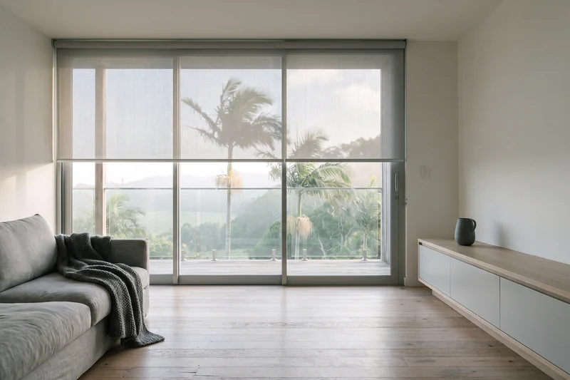 Sunscreen roller blind half lowered across wide glass in a timber coastal house, soft morning light