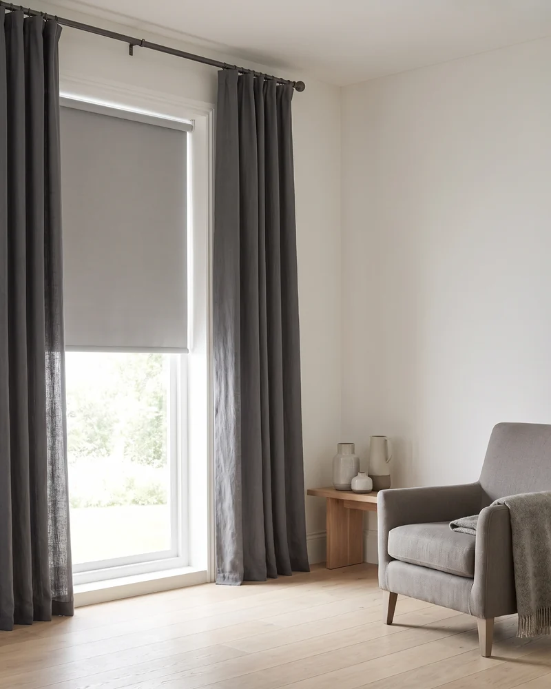 A window dressed with a fabric blind lowered over the glass and long curtain panels at each side
