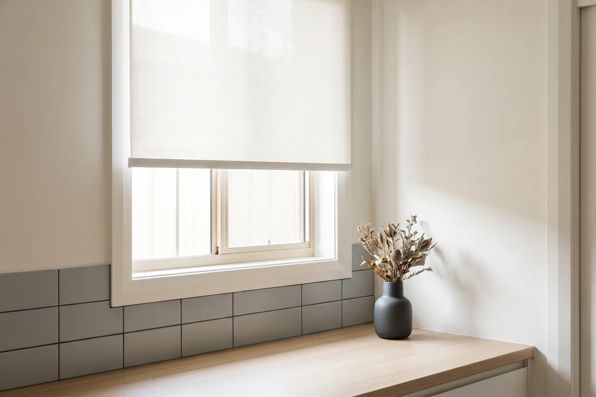 A roller blind half lowered at a kitchen window, sitting clear of the benchtop and tiled splashback