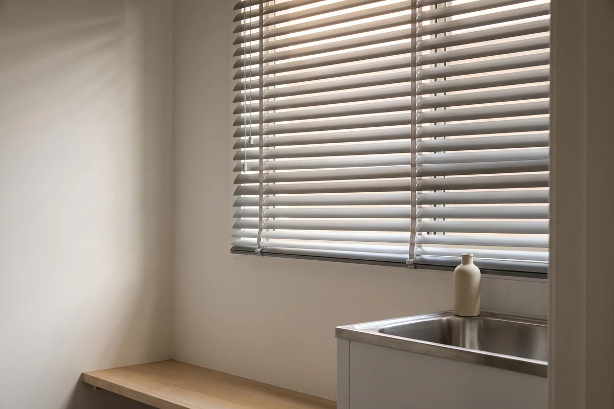 An aluminium venetian blind lowered over a laundry window above a stainless trough, slats tilted part open