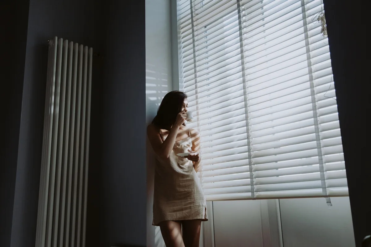 A hand parting venetian blind slats in front of a sunlit window