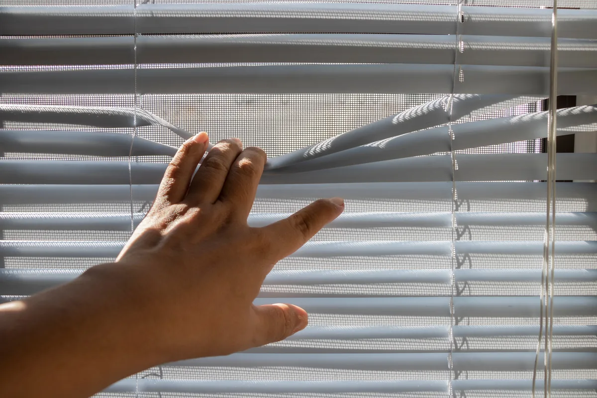A hand parting venetian blind slats in front of a sunlit window