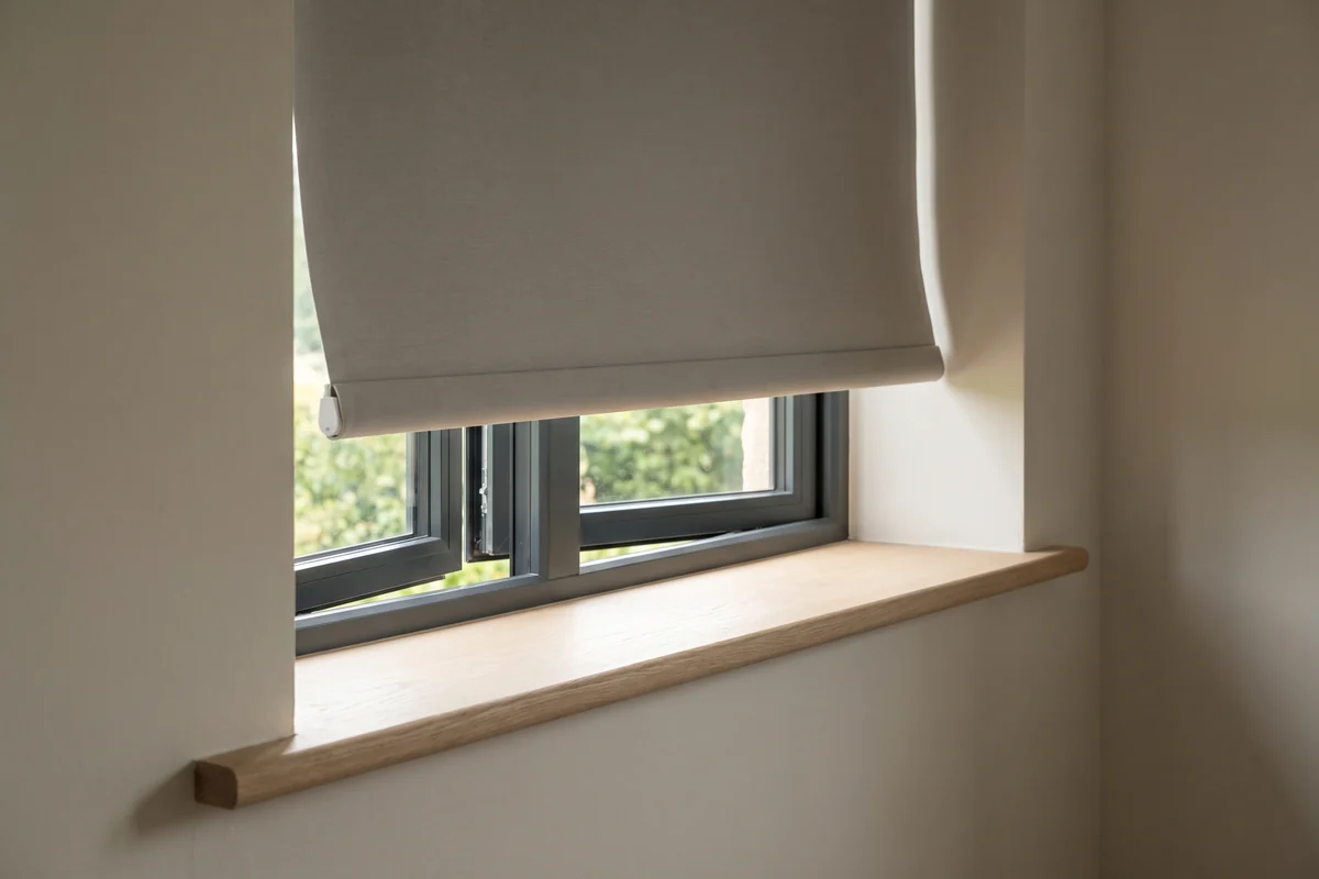 A roller blind at an open window, its bottom rail swung clear of the reveal by the breeze