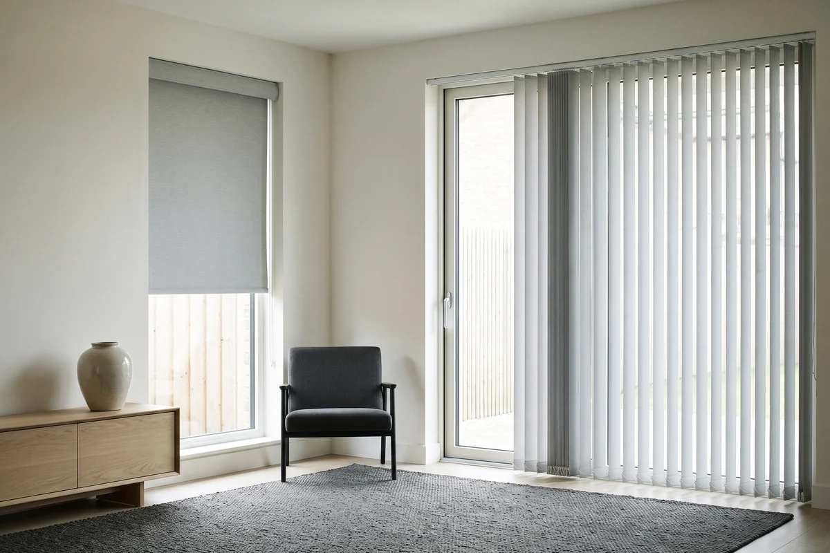 A living room with a roller blind at one window and vertical blinds drawn across the sliding door beside it