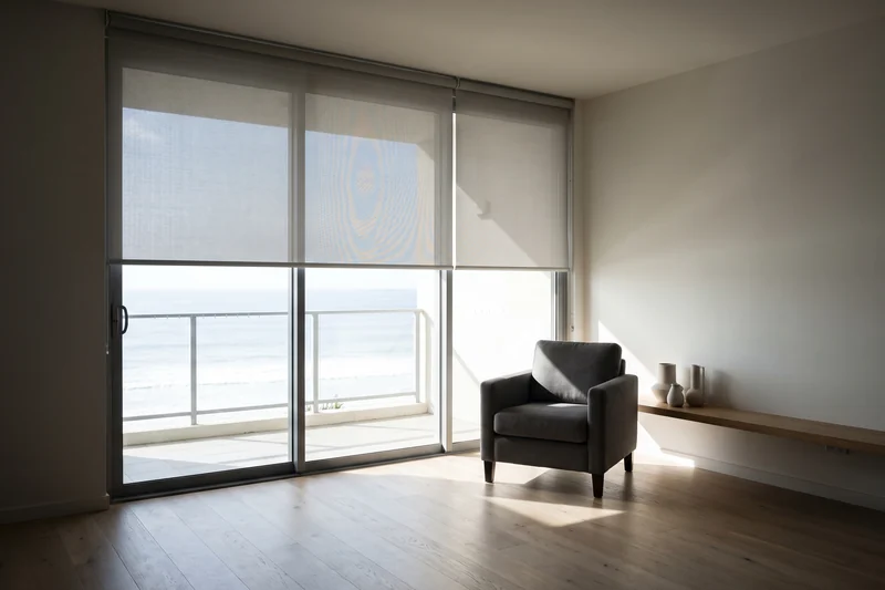 Sunscreen roller blind lowered over a wide glass door with the ocean beyond