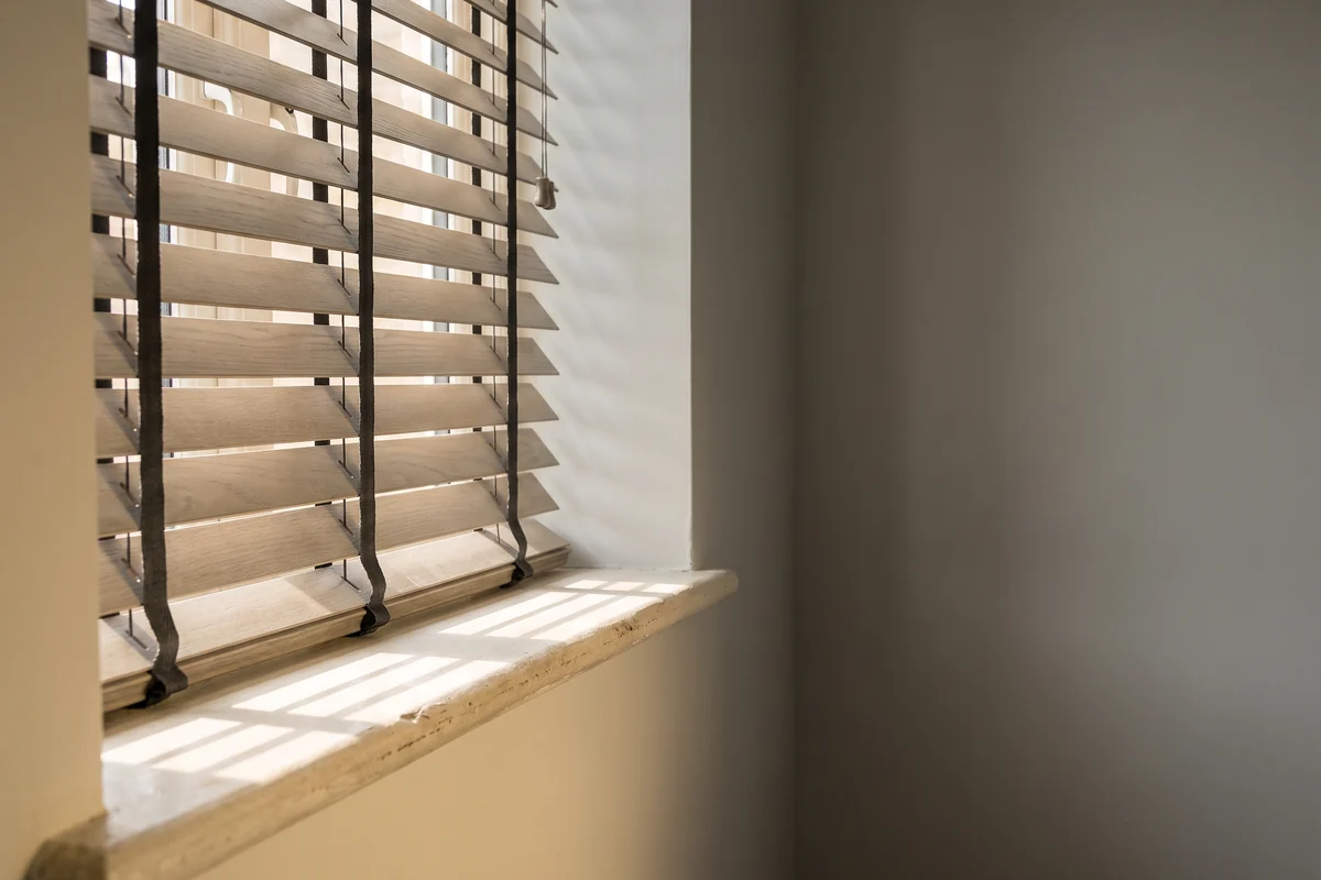 Venetian blind slats tilted part-open, the ladder tape running down each row and daylight striping the sill below